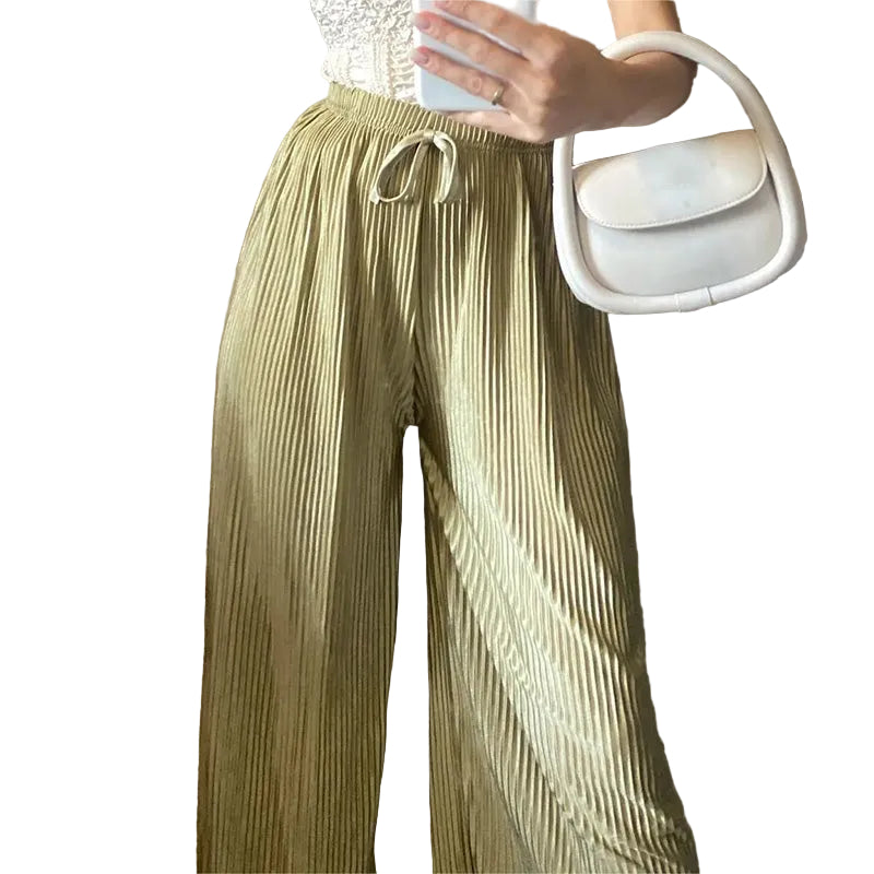 Person wearing pleated green pants holding a white handbag against a brick wall.