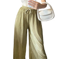 Person wearing pleated green pants holding a white handbag against a brick wall.